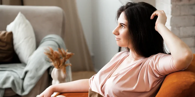 Woman with dark hair sitting thoughtfully on a couch in a living room, leaning back with hand near her head.