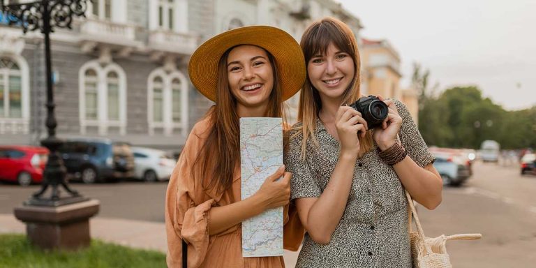Two smiling women travelers holding a map and camera, exploring a city street while managing hormone replacement therapy on vacation.